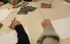 Kilku uczni&oacute;w siedzi przy wsp&oacute;lnym stole, każdy z nich rysuje lub zapisuje coś na kartce. Na stole pomoce naukowe i materiały plastyczne