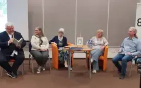 Pięć siedzących na fotelach os&oacute;b, za nimi baner biblioteki i ekran z prezentacją