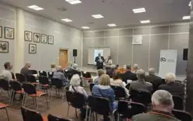 Tyłem do obiektywu siedzi publiczność, przed nią dyrektor biblioteki i prowadząca przemawiają
