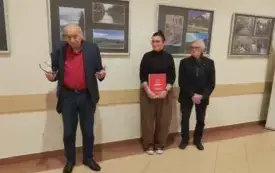 Trzy osoby stoją, jedna z nich przemawia gestykulując, za ich plecami wystawa