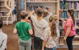 Przedszkolaki przeglądają książki ustawione na bibliotecznych regałach.