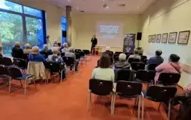 Na pierwszym planie widać uczestnik&oacute;w spotkania. Siedzą na krzesełkach tyłem do aparatu, patrzą na stojącego przed nimi prelegenta. Ten trzyma mikrofon i przemawia do publiczności, za nim wyświetla się strona tytułowa prezentacji. 