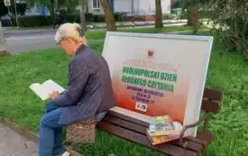 Kobieta siedząca na ławce w parku, czytająca książkę. Obok znajduje się plakat informujący o Og&oacute;lnopolskim Dniu Głośnego Czytania. W tle widać drzewa, trawnik i budynki.