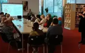 Seniorzy siedzą przy stole i oglądają prezentację podczas spotkania z dietetykiem. Obok nich tłumaczka polskiego języka migowego tłumaczy wypowiedzi prowadzącej.
