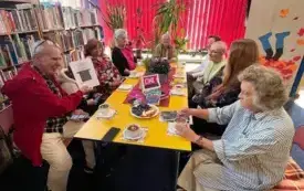 Przy stole siedzi osiem os&oacute;b, mają przed sobą filiżanki i książki.