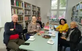 Przy miętowym stole siedzi piątka uczestnik&oacute;w Dyskusyjnego Klubu Książki, trzech mężczyzn i dwie kobiety. Jeden z mężczyzn oraz moderatorka grupy trzymają książki Macieja Siembiedy pt. &bdquo;Gambit&rdquo; w g&oacute;rze. Na stole są książki, słodycze, talerzyki, filiżan