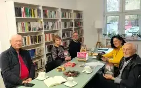 Przy miętowym stole siedzi piątka uczestnik&oacute;w Dyskusyjnego Klubu Książki, trzech mężczyzn i dwie kobiety. Jeden z mężczyzn trzyma książkę Macieja Siembiedy &bdquo;Gambit&rdquo; w g&oacute;rze. Na stole są książki, słodycze, talerzyki, filiżanki, mała sztaluga z logiem Dy