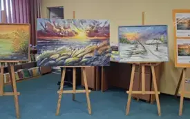 Na sztalugach stoją 3 obrazy oraz plakaty promujące wystawę.