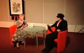 Aktorzy siedzą na pomarańczowych fotelach przy stoliku, inscenizują fragment powieści