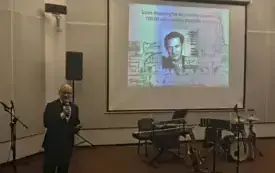 Po lewej stronie znajduje się Rafał Klaus. Z uśmiechem m&oacute;wi do publiczności trzymając mikrofon i patrzy w obiektyw. W tle wyświetlona jest tytułowy slajd jego prezentacji: czas dancing&oacute;w w mieście Gorzowie, 100 lat od urodzin Mariana Klausa. 