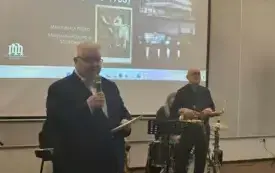 Na pierwszym planie znajduje się Dyrektor Biblioteki. Trzyma mikrofon i przemawia do publiczności otwierając spotkanie. Za nim widać członk&oacute;w zespołu stojących przy instrumentach. Na ścianie wyświetla się tytułowa strona prezentacji prelegentki.