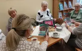 Siedzące przy stole panie, niekt&oacute;re z nich trzymają w rękach książkę, na stole jasna serweta, fioletowy emblemat DKK, bukiet bordowo-zielonych hortensji, filiżanki z napojami, talerzyki, ciasto. W tle jasnoż&oacute;łta ściana i jasnobrązowa p&oacute;łka z książkami.