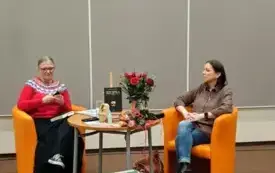 Agnieszka Kopaczyńska-Moskaluk i Danuta Szulczyńska-Miłosz siedzą przy stoliku na kt&oacute;rym znajduje się książka autorki i kwiaty. Przed nimi dekoracja z walizek i maszyny do pisania.