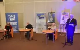 Trzy siedzą przy stolikach. Każdy ma przed sobą mikrofon. W tle osoba prowadząca spotkanie.