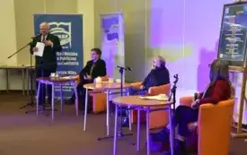 Trzy siedzą przy stolikach. Każdy ma przed sobą mikrofon. W tle osoba prowadząca spotkanie.