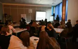 Grupa ludzi siedzi przy stołach. W głębi prelegent.