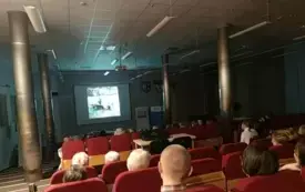 Wewnątrz pomieszczenia panuje p&oacute;łmrok. Publiczność ogląda film wyświetlony na ekranie. Wnętrze biblioteki. Sala audytoryjna.