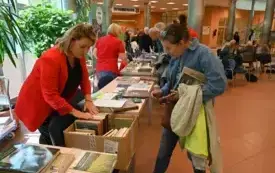 Magdalena Matuszewska rozmawia z czytelnikiem przy stoisku biblioteki. Na stoliku publikacje biblioteki i inne regionalne. Za nimi widać stoliki innych wystawc&oacute;w i zgromadzonych przy nich czytelnik&oacute;w
