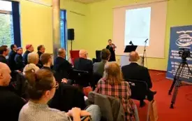 Publiczność siedzi plecami do obiektywu, osoba z przodu gra na skrzypcach, na ekranie wyświetlana jest prezentacja. Kliknięcie powoduje powiększenie zdjęcia.