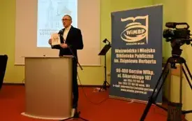 Mężczyzna przy m&oacute;wnicy prezentuje książkę, z tylu wyświetlana jest prezentacja, z prawej strony baner biblioteki. Kliknięcie powoduje powiększenie zdjęcia.