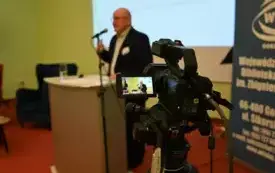 Na pierwszym planie kamera, na drugim mężczyzna przy mikrofonie stoi przy m&oacute;wnicy. Kliknięcie powoduje powiększenie zdjęcia.