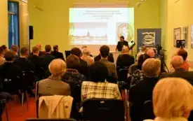 Publiczność siedzi plecami do obiektywu, w tle widać osobę przemawiającą z prezentacją w tle. 