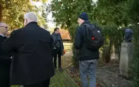 Zdjęcie w plenerze. Kilka os&oacute;b stoi w otoczeniu zieleni przy cokole z popiersiem. 