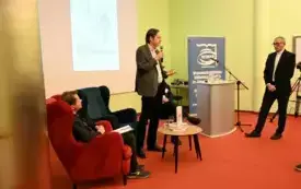 Mężczyzna stoi z mikrofonem i przemawia. Przed nim mały stoliczek, z prawej szara m&oacute;wnica, z lewej dwa fotele, jeden zajęty przez mężczyznę. 