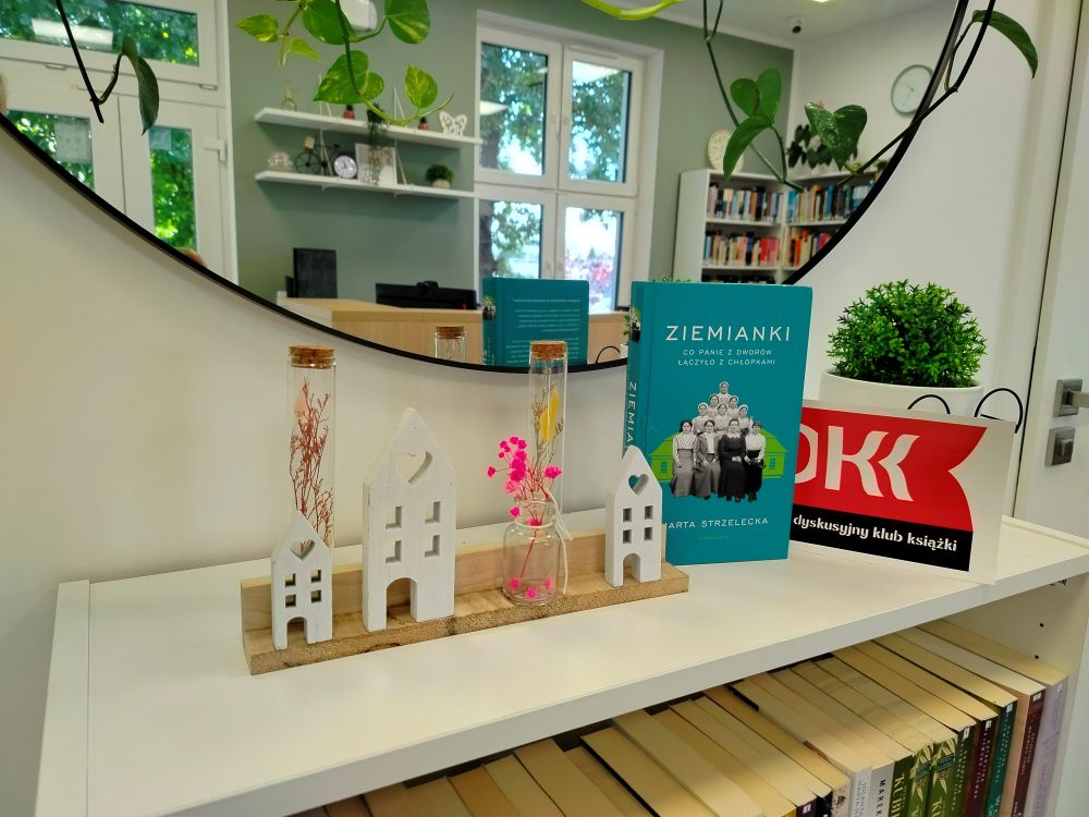 Na małej, białej, komodzie, pod dużym, okrągłym lustrem w&nbsp;czarnej ramie i&nbsp;pnącymi się zielonymi listkami soi książka &bdquo;Ziemianki&rdquo; Marty Strzeleckiej, a&nbsp;przy niej stoją ozdoby:  po lewej stronie od książki zasuszone kwiaty w&nbsp;przezroczystych fiolkach, figurki trzech białych dom&oacute;w na&nbsp;drewnianej, jasnej podstawie. Po prawej stronie logo Dyskusyjnego Klubu Książki oraz&nbsp;biała doniczka w&nbsp;okularach z&nbsp;rośliną w&nbsp;środku. Pod spodem, na&nbsp;p&oacute;łkach komody stoją książki. W lustrze odbijają się drzwi wejściowe do biblioteki oraz&nbsp;miętowa ściana z&nbsp;dwiema białymi p&oacute;łkami, na&nbsp;kt&oacute;rych stoją ozdoby (sztuczne zielone kwiaty, mały rower, świecznik, biała ramka, biały ażurowy motylek, zielony szklany wazonik). Pod p&oacute;łkami widać kawałek jasnej lady i&nbsp;kawałek czarnego monitora.