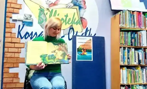 Tytułowe - Kobieta siedzi na krześle i prezentuje treść książki, w tle widać plakat wydarzenia