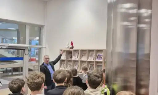 Grupa dzieci stoi tyłem do aparatu w jasnym wnętrzu biblioteki. Pracownik biblioteki – mężczyzna, wskazuje na półkę z czasopismami, o których opowiada dzieciom. Po prawej stronie pomieszczenia znajdują się srebrne filary oraz jasny regał z czasopismami. T