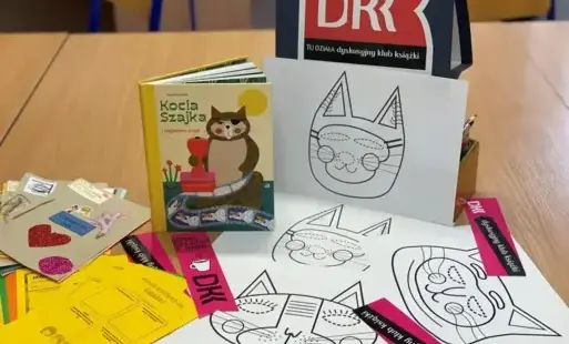 Zdjęcie startowe – przedstawia książkę A. Romaniuk „Kocia Szajka i zaginione bajki”, napis DKK, zakładki DKK, dzienniczki DKK, karty oceny oraz kartki z kocimi maskami.