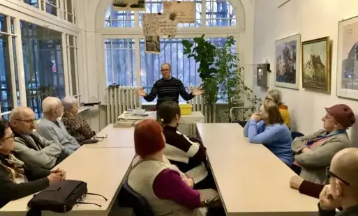 Spotkanie grupowe w jasnej sali. Przy dwóch długich stołach siedzi kilkanaście osób ubrane w swetry i bluzy w stonowanych kolorach; część ma siwe włosy, jedna osoba z przodu ma intensywnie rude włosy, inna malinową czapkę. W centrum między stołami stoi st
