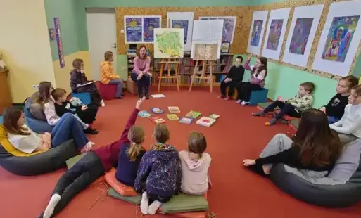 Dzieci wraz z bibliotekarką siedzą na dywanie w kółeczku, przed nimi leżą książki z baśniami i legendami polskimi. W tle widać mapę Polski oraz krzyżówkę. Uczestnicy zajęć rozmawiają.