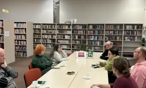 Przy długim stole w Wypożyczalni Głównej siedzą uczestnicy Dyskusyjnego Klubu Książki. Przed nimi leżą egzemplarze książki Oida autorstwa Marta Michalak. Na stole stoi ramka z logo Z DKK, stoją filiżanki z kawą i herbatą, na talerzyku leżą ciastka. W tle 