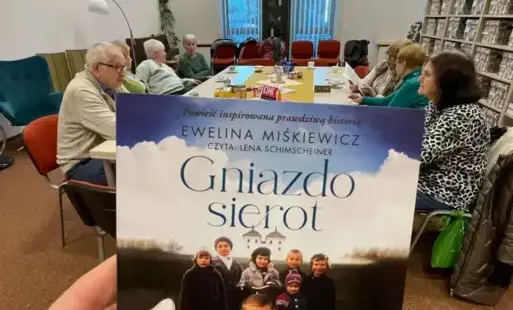 Na pierwszym planie opakowanie od audiobooka, w tle długi stół i siedzące osoby dorosłe.