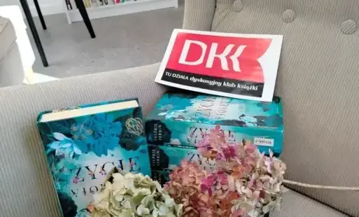 1.	Cztery egzemplarze książki „Życie Violette” Valerie Perrin leżą na beżowym fotelu. Piąty egzemplarz stoi obok. Przy książkach leżą zasuszone hortensje. Na książkach leży logo Dyskusyjnego Klubu Książki. W tle widać biały regał z książkami, krzesło i pu