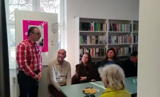 Na zdjęciu widać prowadzącego spotkanie Zbigniewa Rudzińskiego oraz część uczestników spotkania. W tle widać okno, białą tablicę z plakatami i białe ragały z książkami. Na środku stoi stół. Na stole leży talerz ze słodyczami, okulary.