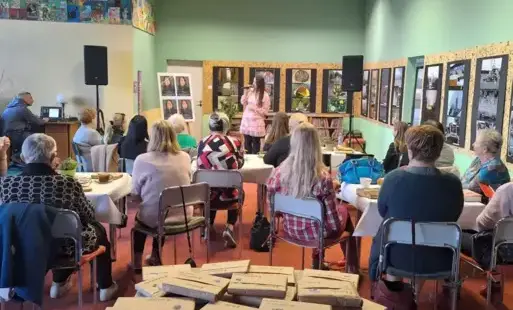 W sali bibliotecznej przy stolikach nakrytych białymi obrusami siedzą kobiety, piją kawę lub herbatę i słuchają koncertu. Przed nimi stoi wokalistka w różowej sukience i śpiewa. W tle wisi wystawa fotografii. Za publicznością znajduje się stolik z zapakow
