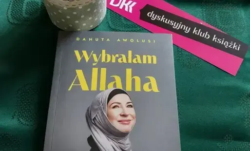 Na zielonym obrusie książka ,, Wybrałam Allaha. Polki, które przeszły na islam” autorstwa Danuty Awolusi, emblemat Dyskusyjny Klub Książki i wiosenne bazie w małym wazoniku.