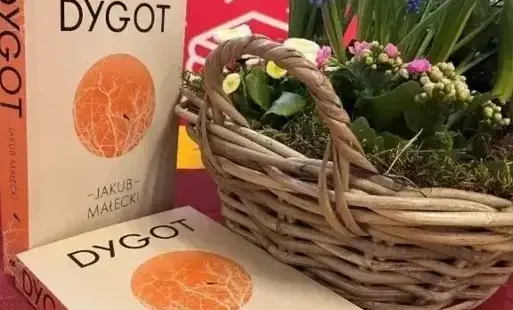 na stole leżą książki, w tle koszyk z kwiatami i plakat DKK