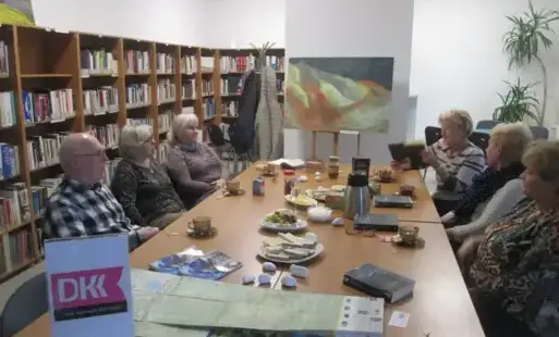 Sześć osób siedzi przy szerokim stole kawowym z logo Dyskusyjnego Klubu Książki, jedna z nich podnosi książkę i coś z niej czyta z uśmiechem. Na blacie między filiżankami i talerzykami leży mapa i białe kamienie. W tle abstrakcyjny obraz w kolorach czarny