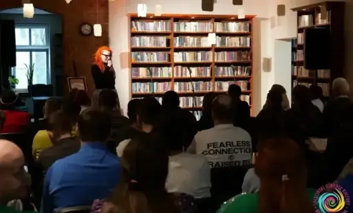 Kobieta z mikrofonem naprzeciwko siedzących osób w pomieszczeniu biblioteki.