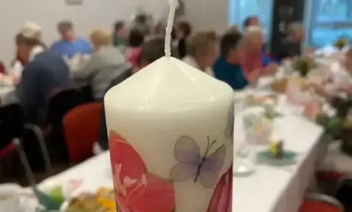 Na pierwszym planie wielkanocna świeca, a w rozmytym tle przy nakrytych stołach siedzą dorosłe osoby.