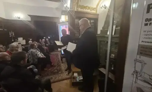 Grupa ludzi siedzących w pomieszczeniu, słuchających mężczyzny stojącego z przodu i trzymającego kartkę. Po lewej stronie widać kilka rzędów osób siedzących blisko siebie; są to głównie osoby starsze. Z przodu, bliżej prawej strony kadru, stoi  mężczyzna.