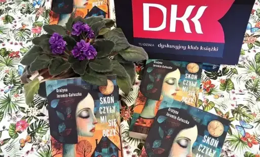 Na stole leżą trzy omawiane książki. W środku stoi doniczkowy fiołek z fioletowymi kwiatami. Obok jest tabliczka z logo DKK.