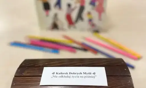 Drewniany, kuferek na stoliku z napisem na małej karteczce: Kuferek Dobrych Myśli. Nie odkładaj życia na później. W tle stoi książka i leżą kredki.