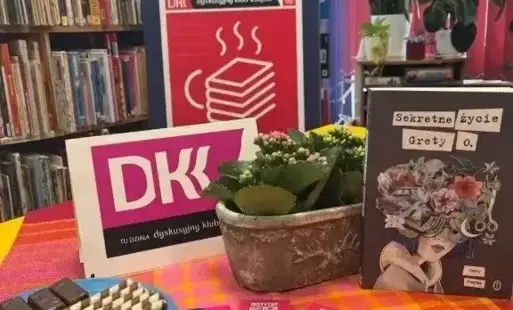 Tytułowe- na stole postawiona książka, doniczka z kwiatkiem i talerzyk z ciastkami
