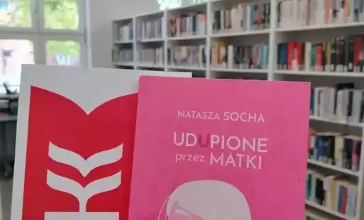 Widać rękę trzymającą książkę „Udupione przez matki” Nataszy Sochy i logo Dyskusyjnego Klubu Książki. W tle są okna z tyłu, po prawej stronie ciąg białych regałów z książkami. Widać kawałek stołu, na którym są książki i parapet z roślinami. Pomieszczen