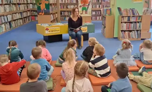 Dzieci siedzą na kolorowych poduszkach na dywanie przed bibliotekarką, która czyta im książeczkę o życiu Hansa Christiana Andersena. W tle znajdują się regały z książkami.